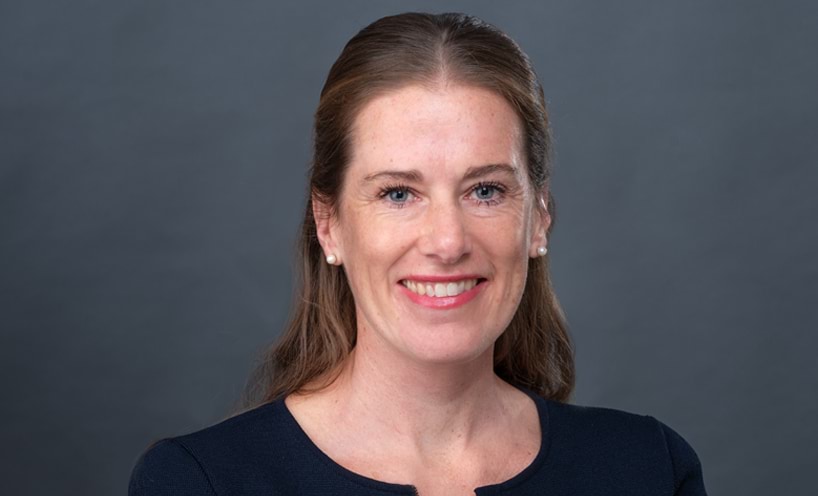 Ella has her long light brown hair half tied up and is wearing a dark blazer.