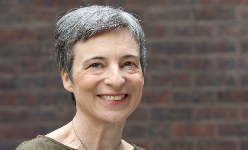 A head and shoulders portrait of Llewellyn, who is sitting in front of a dark brick wall. Llewellyn has short silver hair, fair skin, green-grey eyes and is smiling warmly towards the camera.
